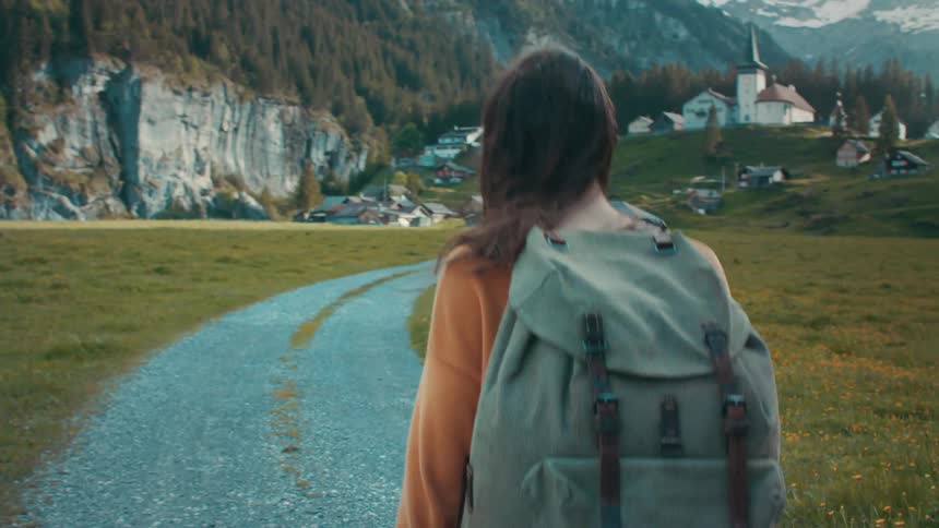 Woman with brown wavy hair orange sweater and backpack walks on country road leading to village in mountainous area