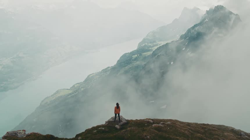 An aerial view of fog rolling over mountains and a woman standing on the edge looking over the valley