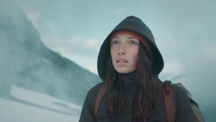 Woman with brown wavy hair green coat and backpack stands and looks into space in snowy mountain