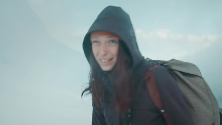 A teenage girl wearing a hooded jacket and a big backpack is walking near mountains as snow blows in the air