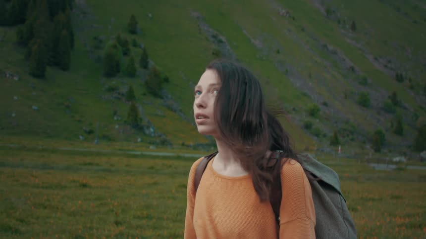 Woman with brown hair orange sweater and backpack walks in the green valley