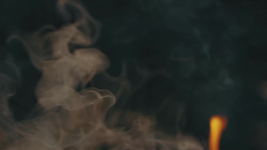 Smoke rises from barrel as flame burns on ground nearby both contrasting against darkness
