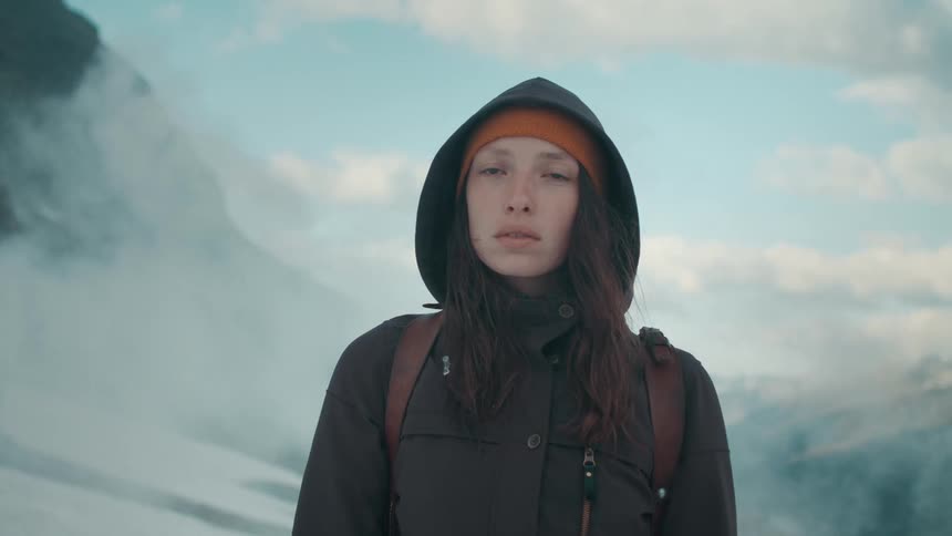 A young woman wearing a winter coat and backpack stands in the mountains as clouds of fog pass behind her