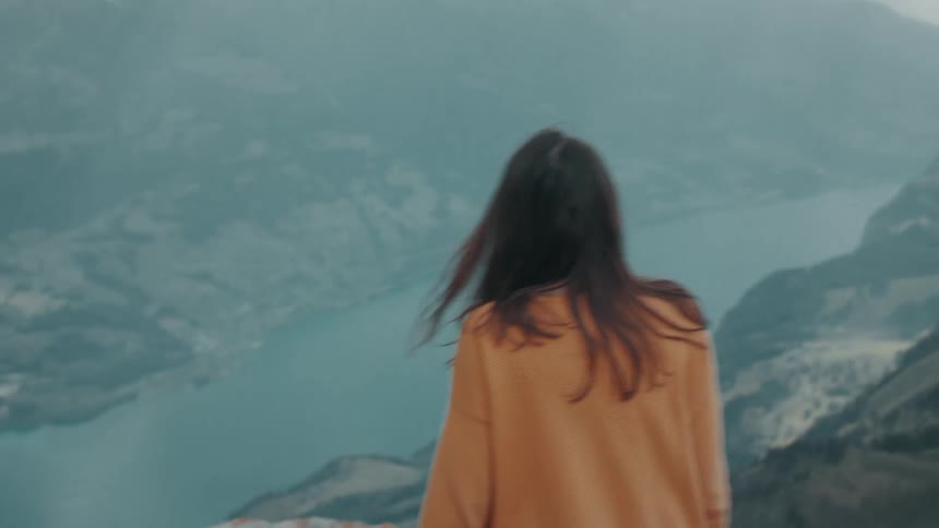 Woman with brown wavy hair and orange sweater takes off backpack and stands at the edge of high mountain overlooking lake and distant mountains