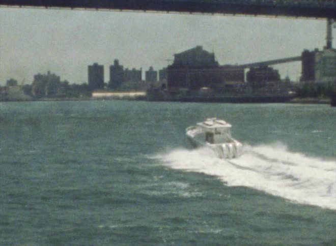 A small motorboat sails under a suspension bridge and the big city buildings and skyscrapers are in the distance