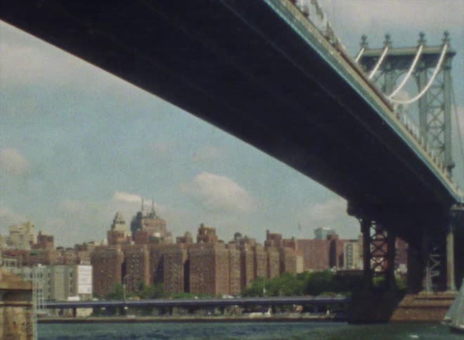 Vehicles drive past downtown along a river with a long bridge overhead