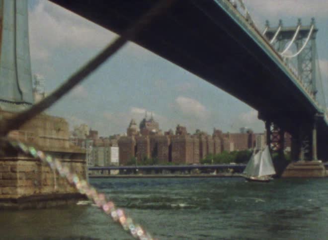 A sailboat sailing under a bridge next to a big city
