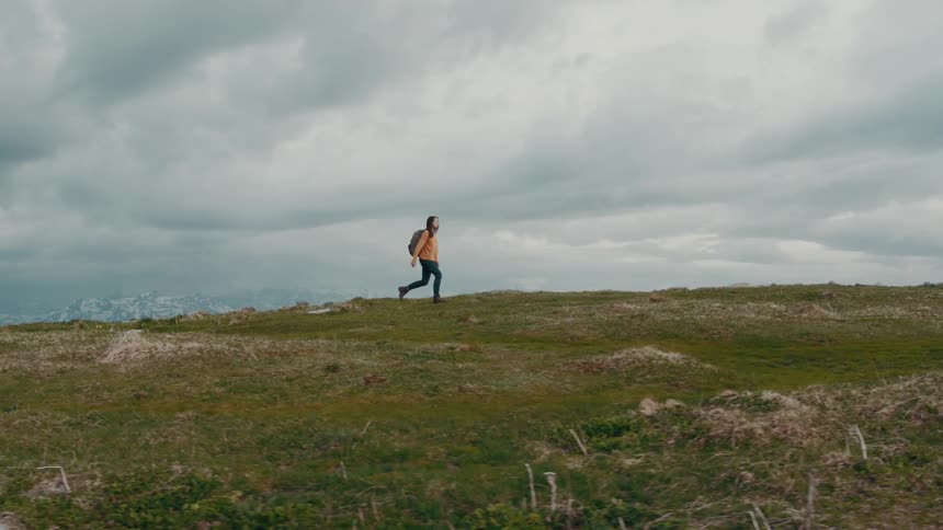 Woman with orange sweatshirt jeans and backpack hikes in the mountains on a cloudy day