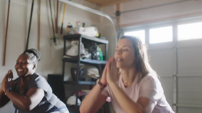 A pair of women chat while doing squats together in a garage during the day