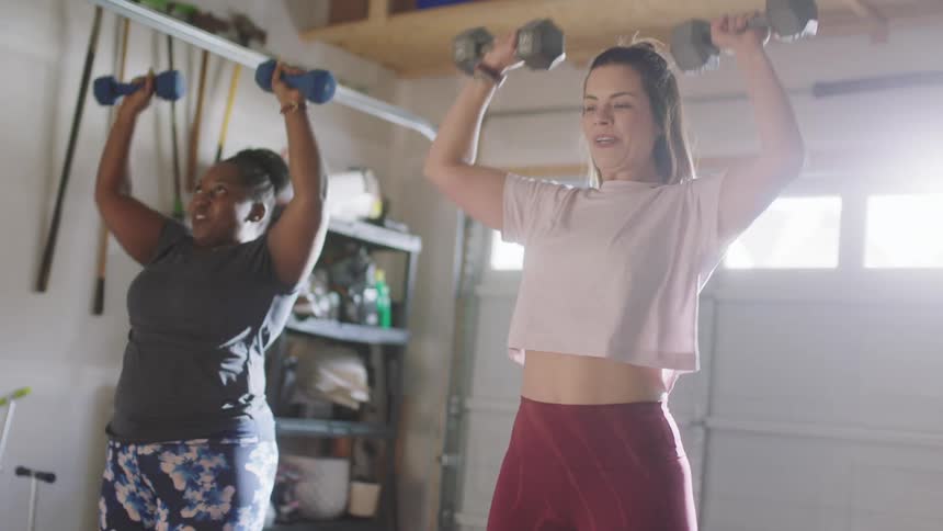 Two women lifting hand weights over their heads while smiling in a garage
