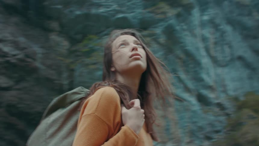 A teenage girl wearing a big backpack is walking near mountains looking around as she holds the strap of her backpack