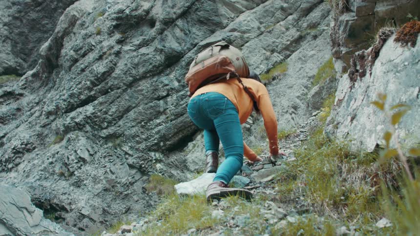 Woman with a pair of boots jeans orange sweatshirt and backpack climbs up steep mountain
