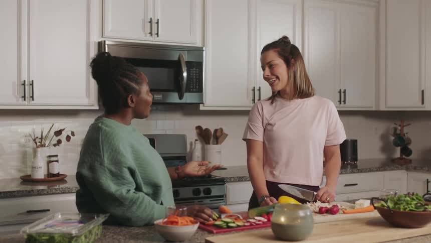 A woman with short hair holding a knife smiling laughing and hugging another women in the kitchen