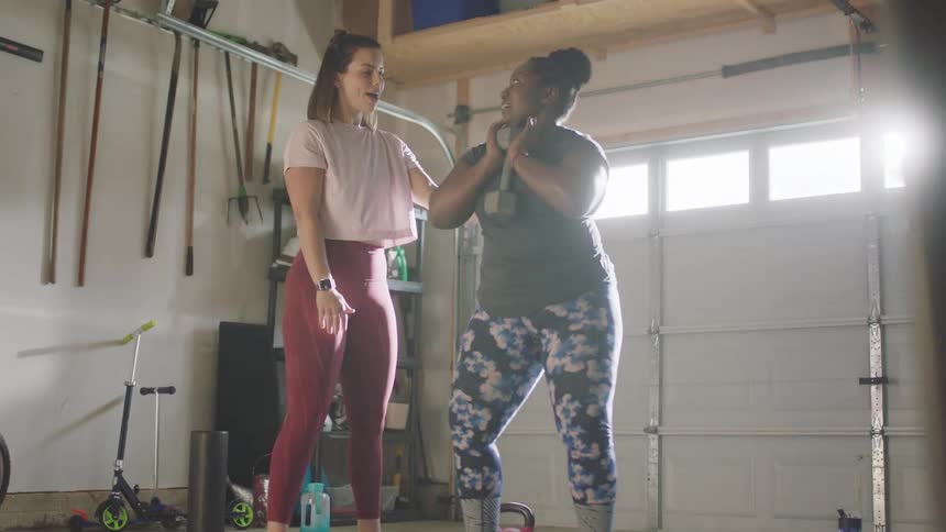 Woman in red yoga pants and pink t shirt helps woman with bun black t shirt and yoga pants do goblet squat in room