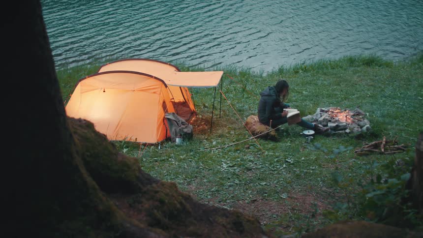 Female camper sits on shore near campfire with journal
