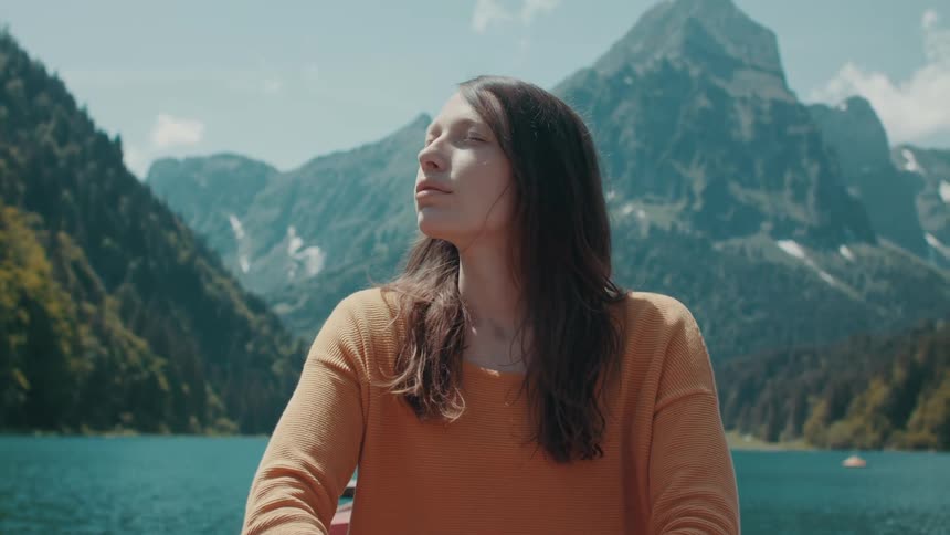 Woman with brown hair and orange sweater paddles canoe along the lake and looks at high mountains