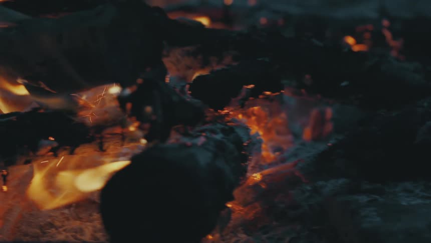 An up close of coals of a fire with small flames burning wood