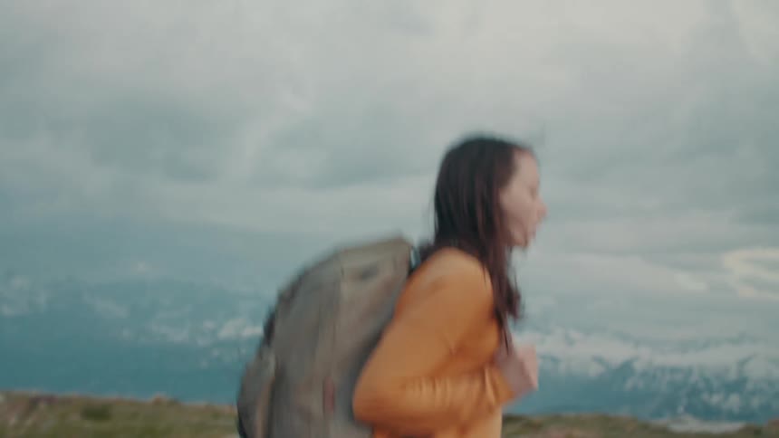 A teenage girl wearing a big backpack is walking across high ground and there are mountains in the distance