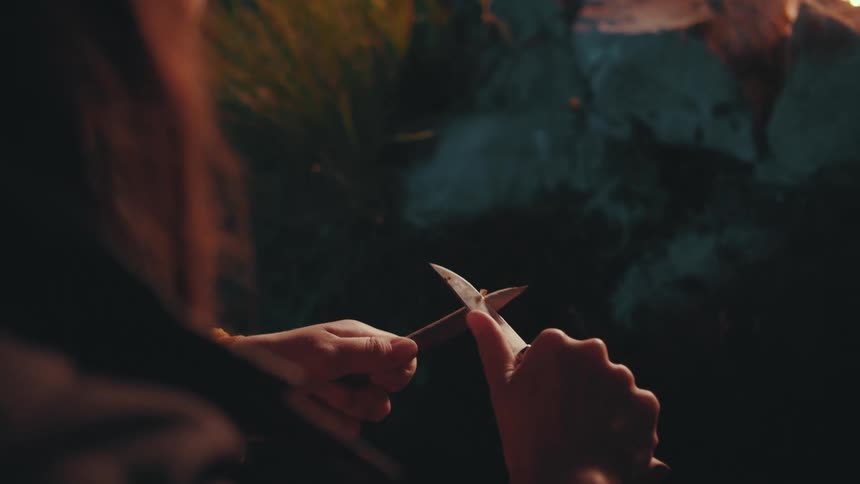 A girl is sharpening a stick with a pocketknife near a campfire