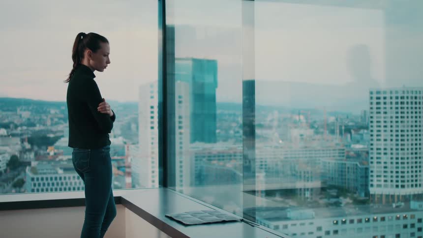 A woman stand and looks out of an office window in a downtown area of a city