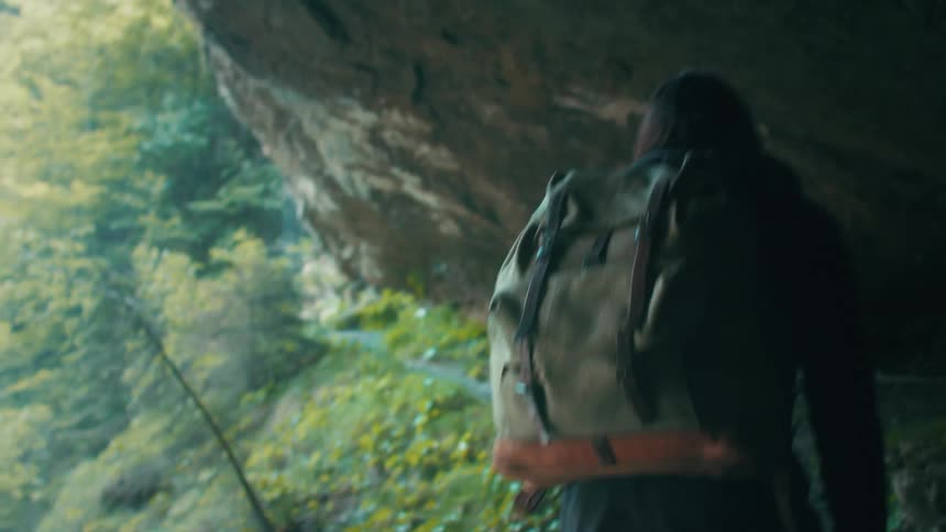 A woman hikes along a narrow dirt trail under a cliff in the wilderness during the day