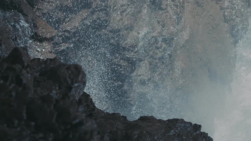 Water is flowing down rocks on a mountain cliff and splashing into the air and there is a stream below