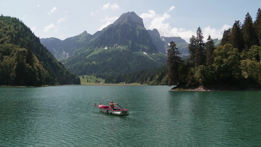 A woman rows a boat along a lake at the bottom of a mountain valley covered in forest