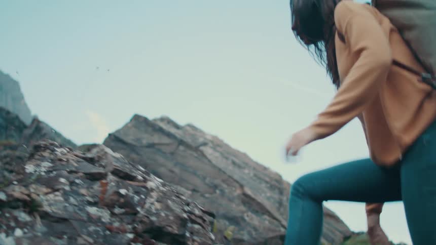 Woman with brown hair orange sweater jeans and backpack climbs up steep mountain