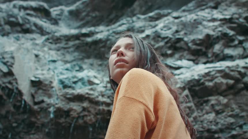 A teenage girl with long dark hair is standing next to a mountain cliff as water trickles down
