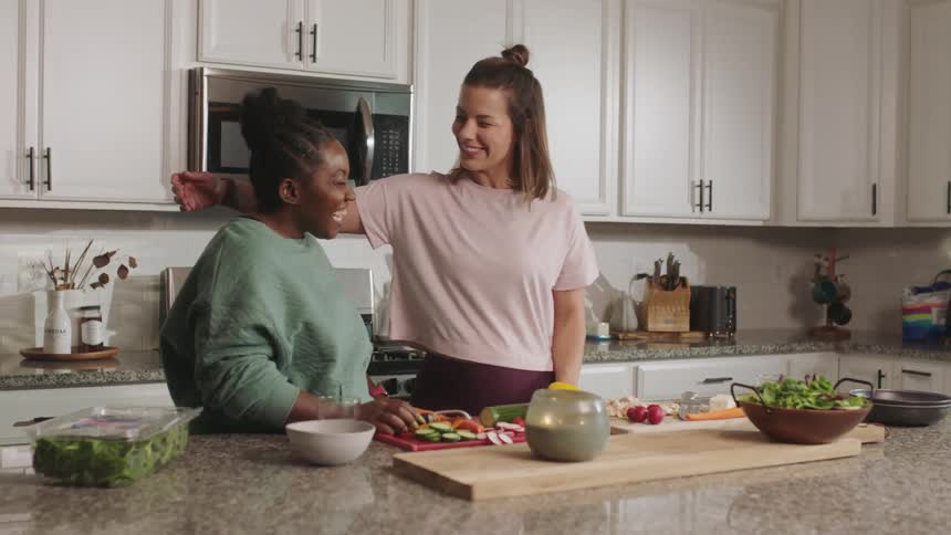 Two women are standing at a kitchen counter cutting vegetables and talking and smiling as one woman hugs the other