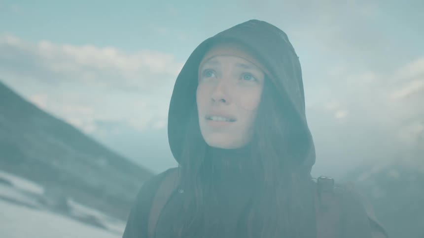 A woman wearing a black coat with her hood up looking up towards the top of a mountain with mist rolling across her face