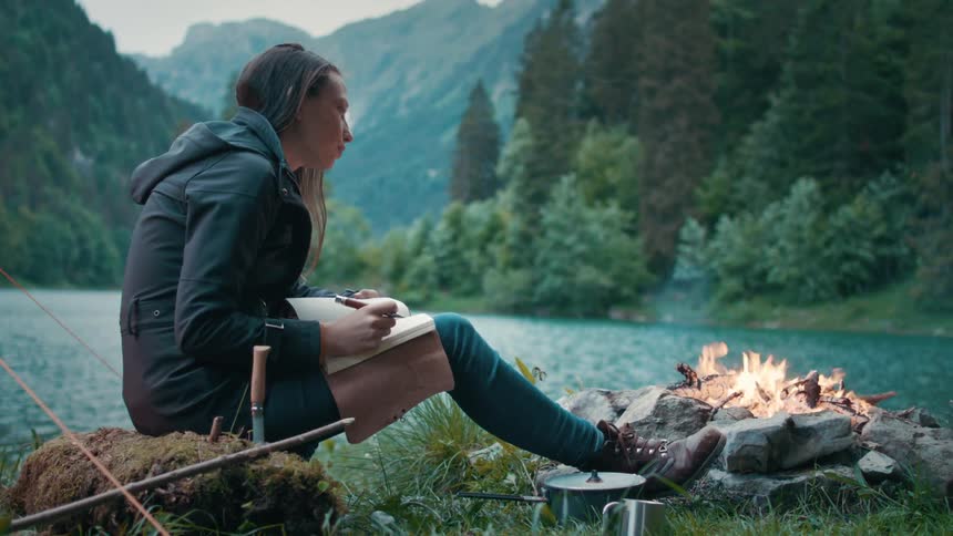 Woman writes in notebook while sitting near campfire on shore across water from forested mountains
