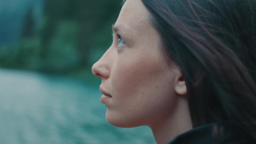 A young woman stands next to a river with her eyes closed before opening them and looking upwards