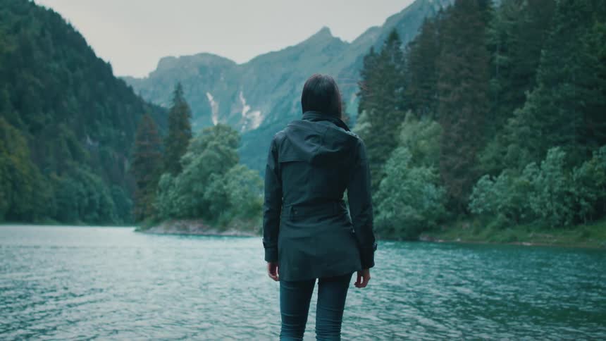 A woman stands on the shore of a river running through a verdant mountain valley during the day