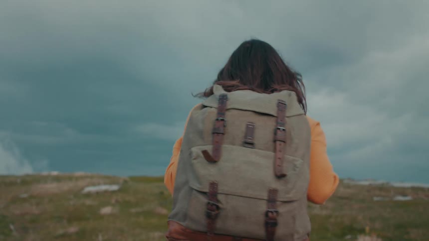 A teenage girl wearing a big backpack is hiking up a hill under a cloudy sky