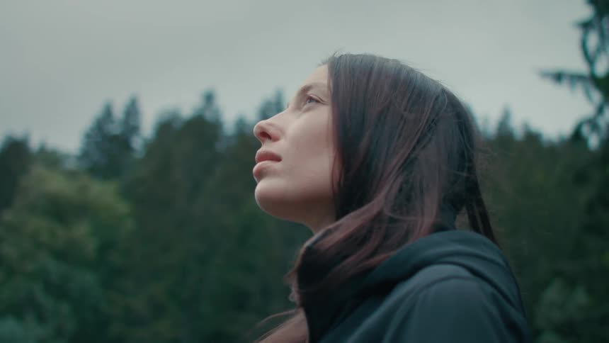 A young woman looks thoughtfully at the sky while standing in the wilderness on a windy day