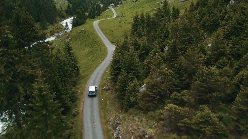 An aerial view of a car driving down on a road through tall pine trees and a river running near the road
