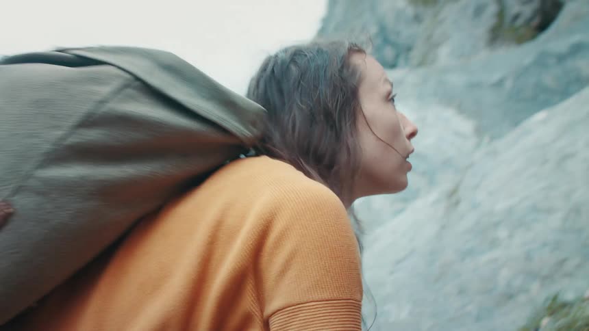 Woman with brown hair orange sweater jeans and backpack climbs up steep mountain