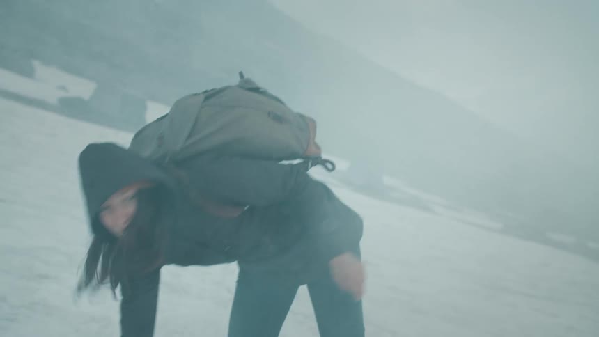A young woman stumbles and looks around in confusion while attempting to ascend a foggy mountain slope covered in snow
