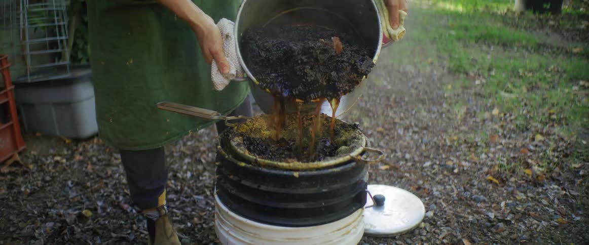 A man pours a pot of sludgy liquid into a sieve over a barrel in his backyard