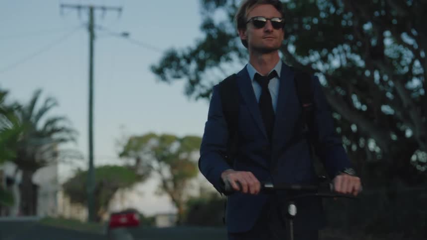 Man wearing sunglasses and dark blue suit rides electric scooter on the street