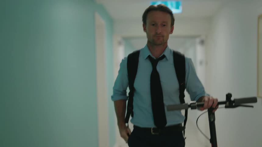 A man looks at his phone as he walks his scooter down a hallway