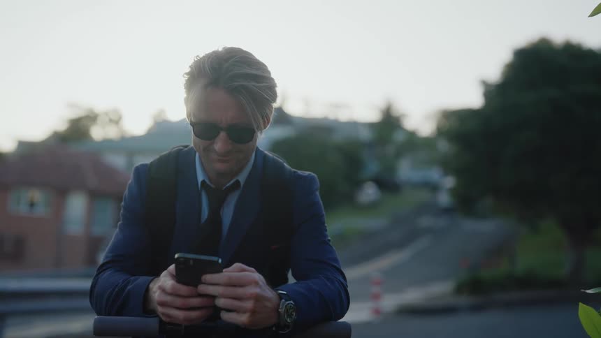 A man wearing sunglasses and a suit is outside using his cell phone as cars travel on the neighborhood road in the distance