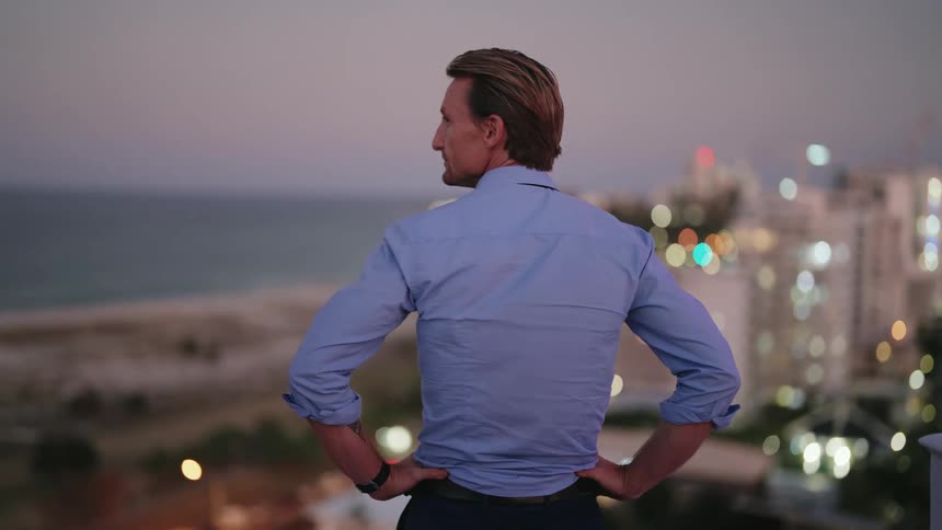 A man is standing outside with his hands on his hips overlooking buildings on a street along the coast and a beach