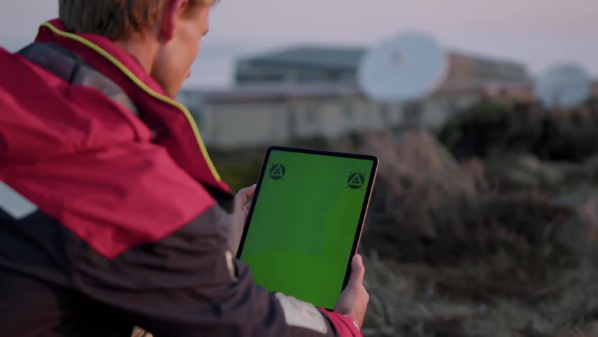A man is sitting outside holding a tablet with a green screen