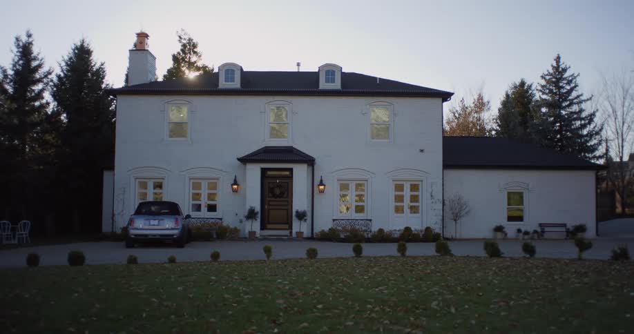 An automobile is parked in front of a large white estate house as the sun sets behind the trees under a clear sky