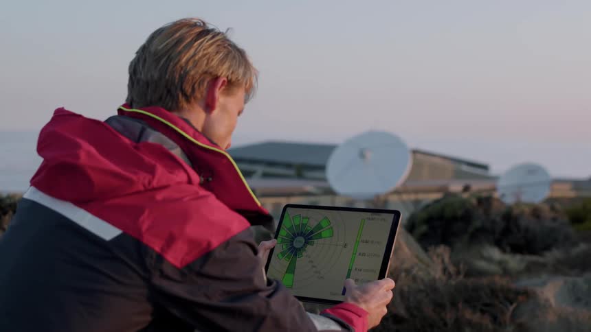 A man is sitting outside using a tablet computer and there are satellite dishes in the distance