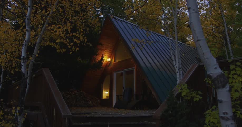 A house in the woods with chairs on the porch is surrounded by birch trees with leaves in fall colors and the lights come on inside