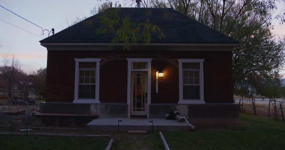The porch light of a small house is on and another light comes on inside