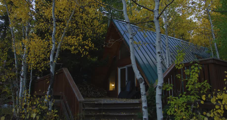 A house in the woods with chairs on the porch is surrounded by birch trees with leaves in fall colors and a light is on inside and another light flashes